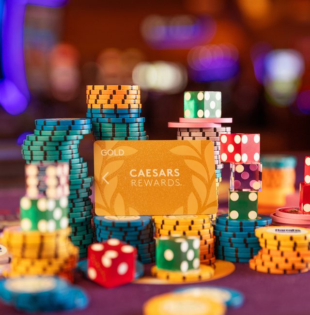 A collection of colorful casino chips stacked around a Caesar's Rewards membership card on a gaming table. The background features blurred lights typical of a casino atmosphere.